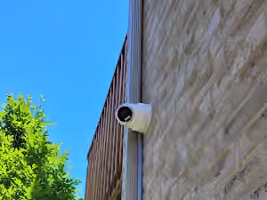 White outdoor security camera on an exterior brick wall beside a downspout.