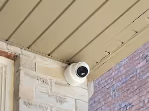 White outdoor security camera mounted beneath a porch soffit beside a brick wall.