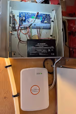 Open alarm control panel with wiring and backup battery above a white communications unit.